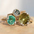 Three gold rings with large round gemstones in green, blue, and olive-yellow tones, each accented with small clear stones, displayed on a textured stone surface.