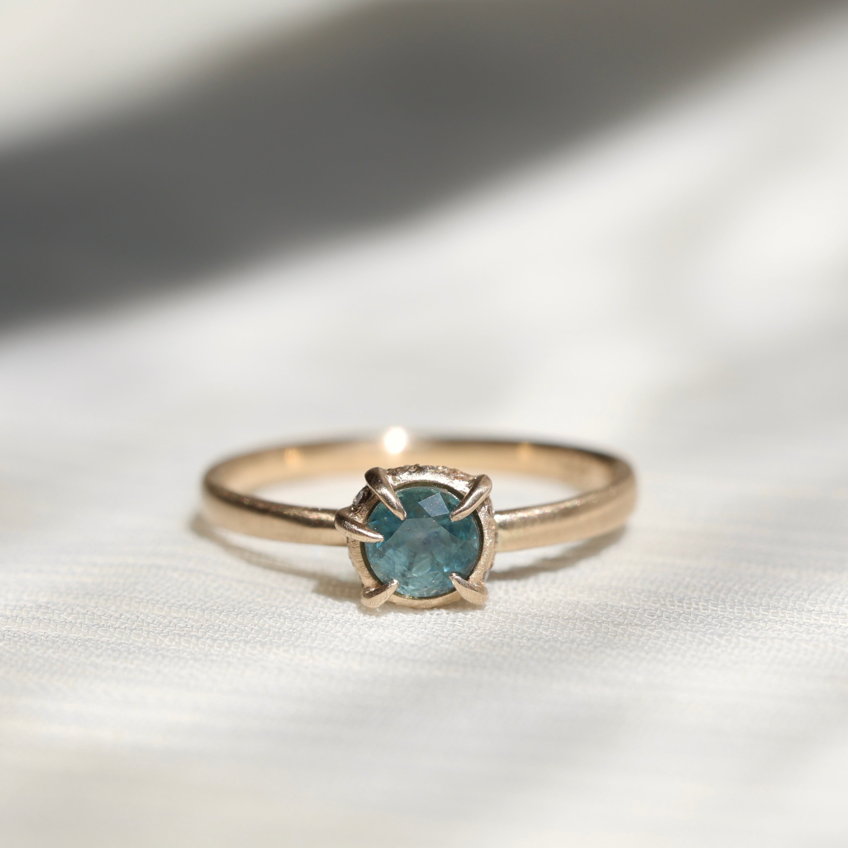 14k yellow gold vanity ring with  a round teal blue Montana sapphire and a halo of small diamonds around the setting, on a white fabric background. 