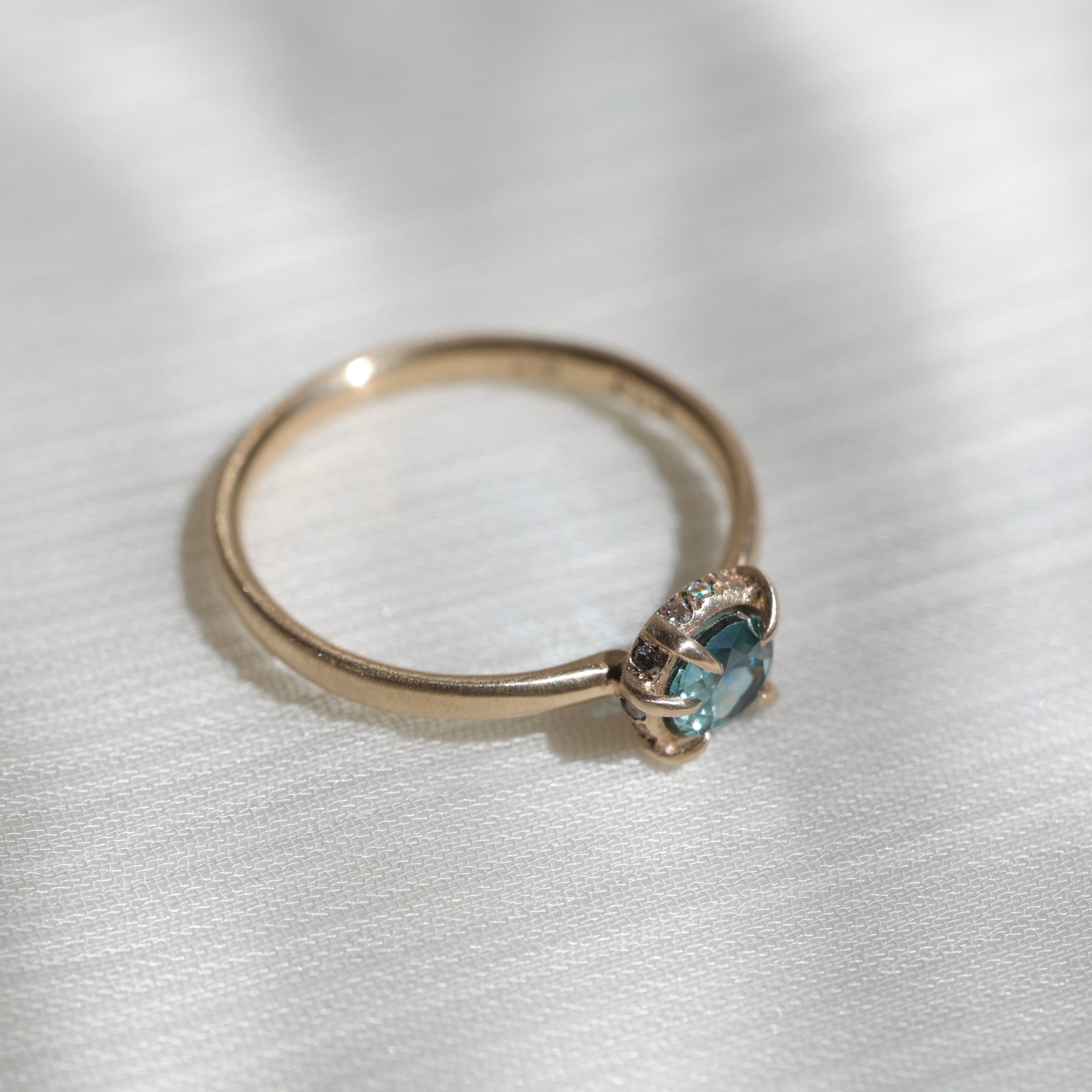 14k yellow gold vanity ring with  a round teal blue Montana sapphire and a halo of small diamonds around the setting, on a white fabric background, side view. 