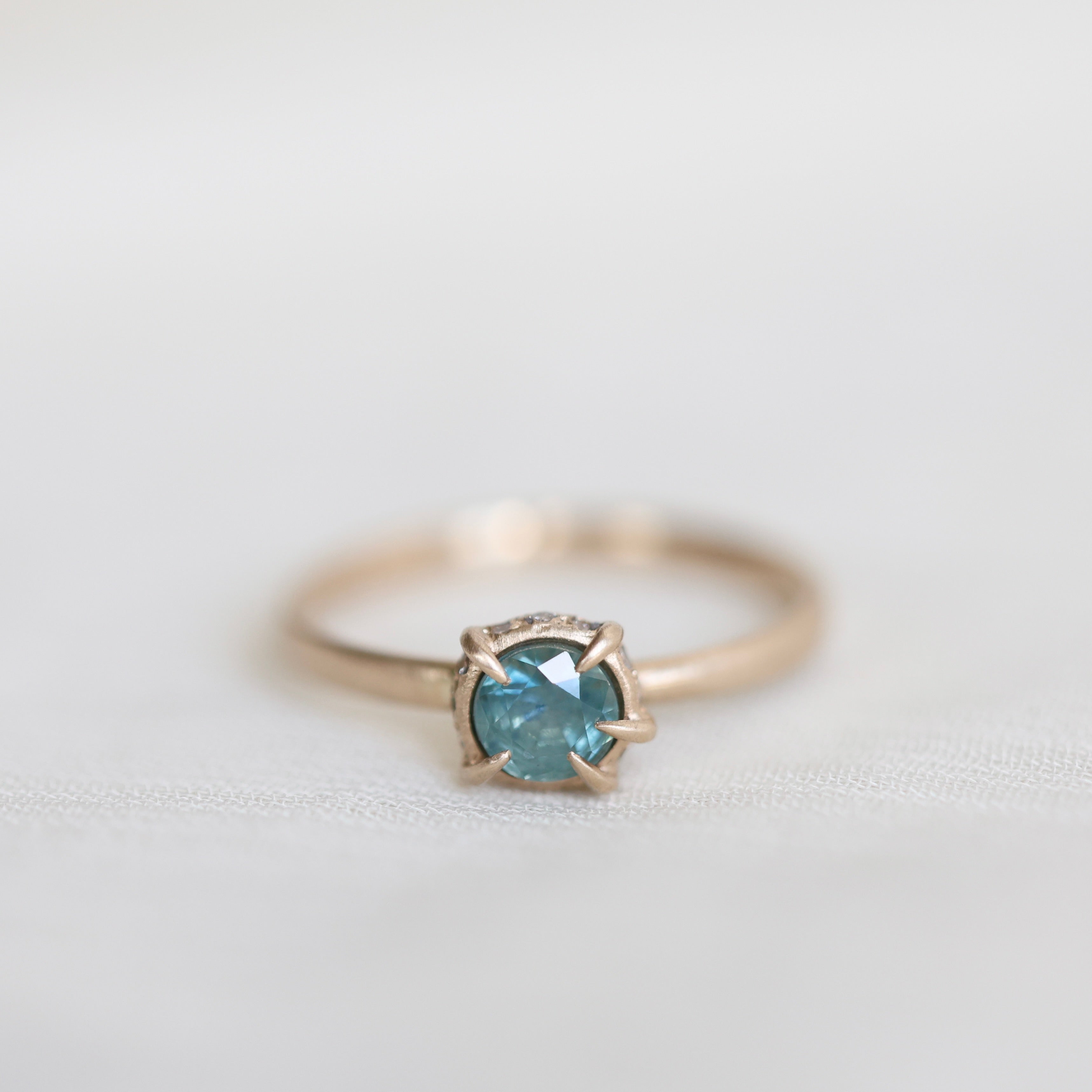14k yellow gold vanity ring with  a round teal blue Montana sapphire and a halo of small diamonds around the setting, on a white fabric background. 