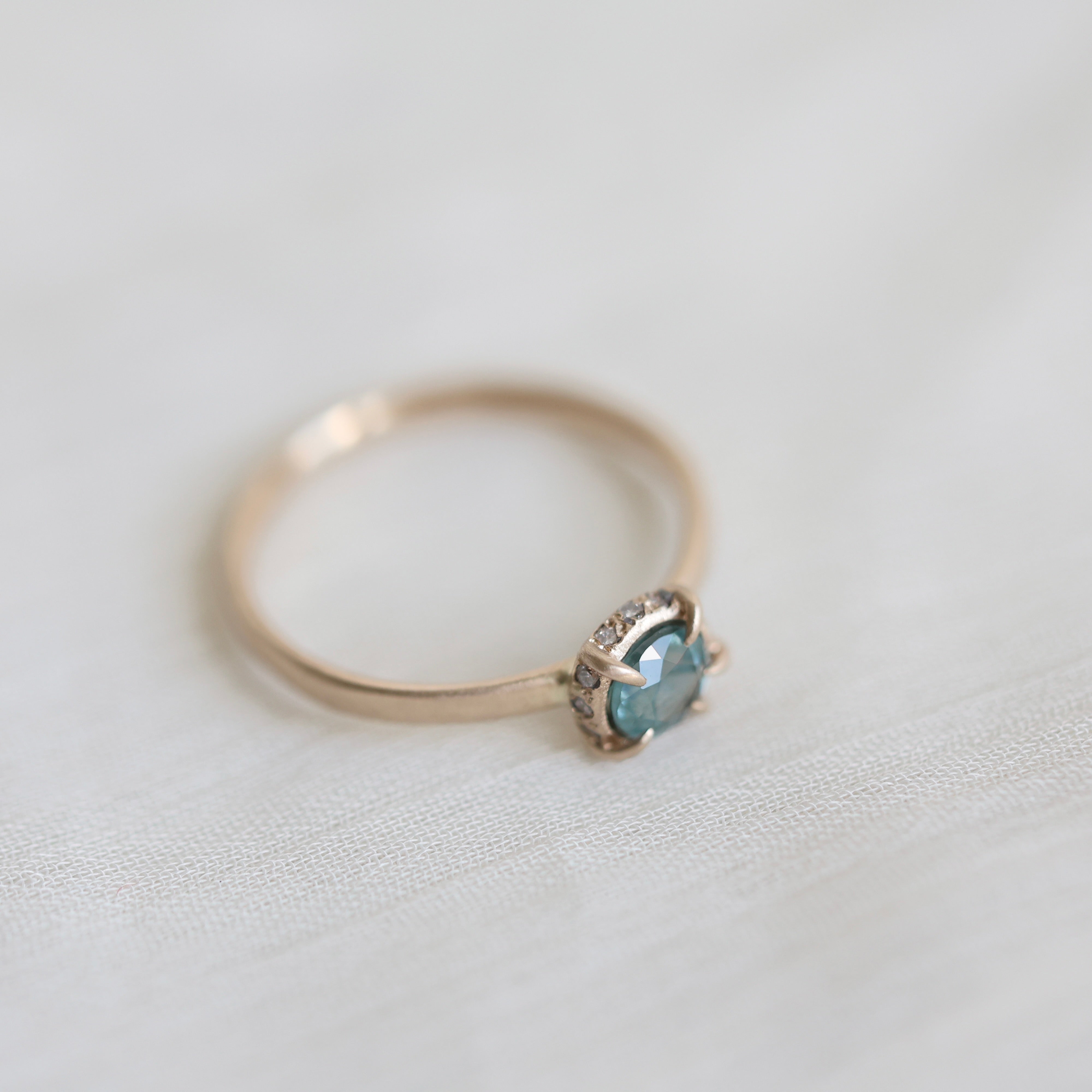 14k yellow gold vanity ring with  a round teal blue Montana sapphire and a halo of small diamonds around the setting, on a white fabric background, side view. 