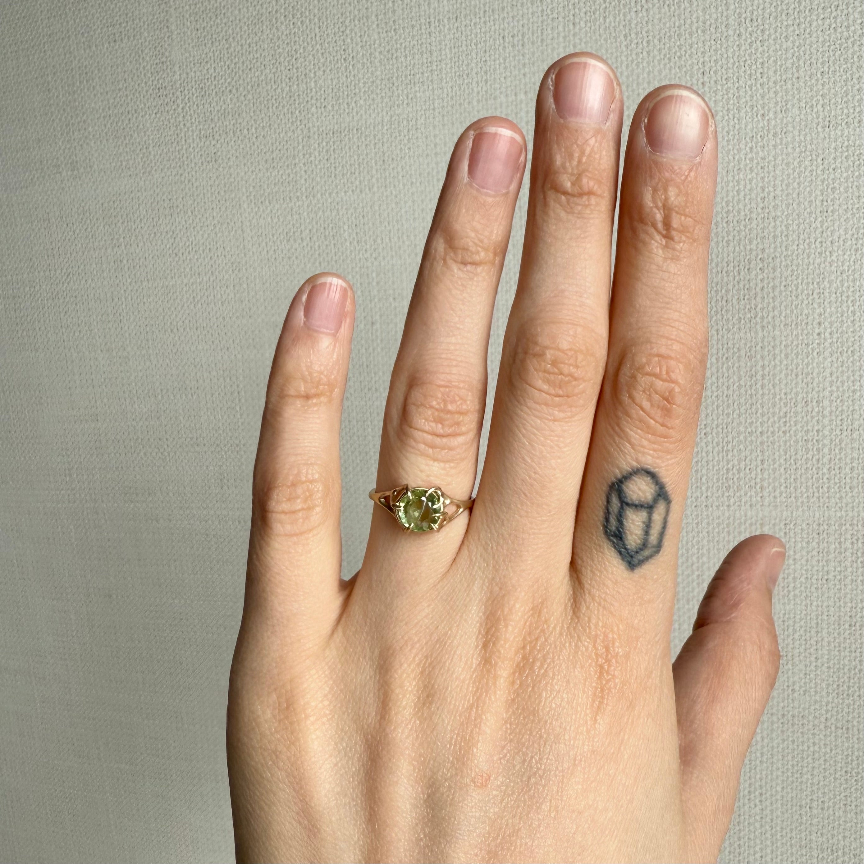 Hand wearing a gold ring with a green gemstone on a neutral background
