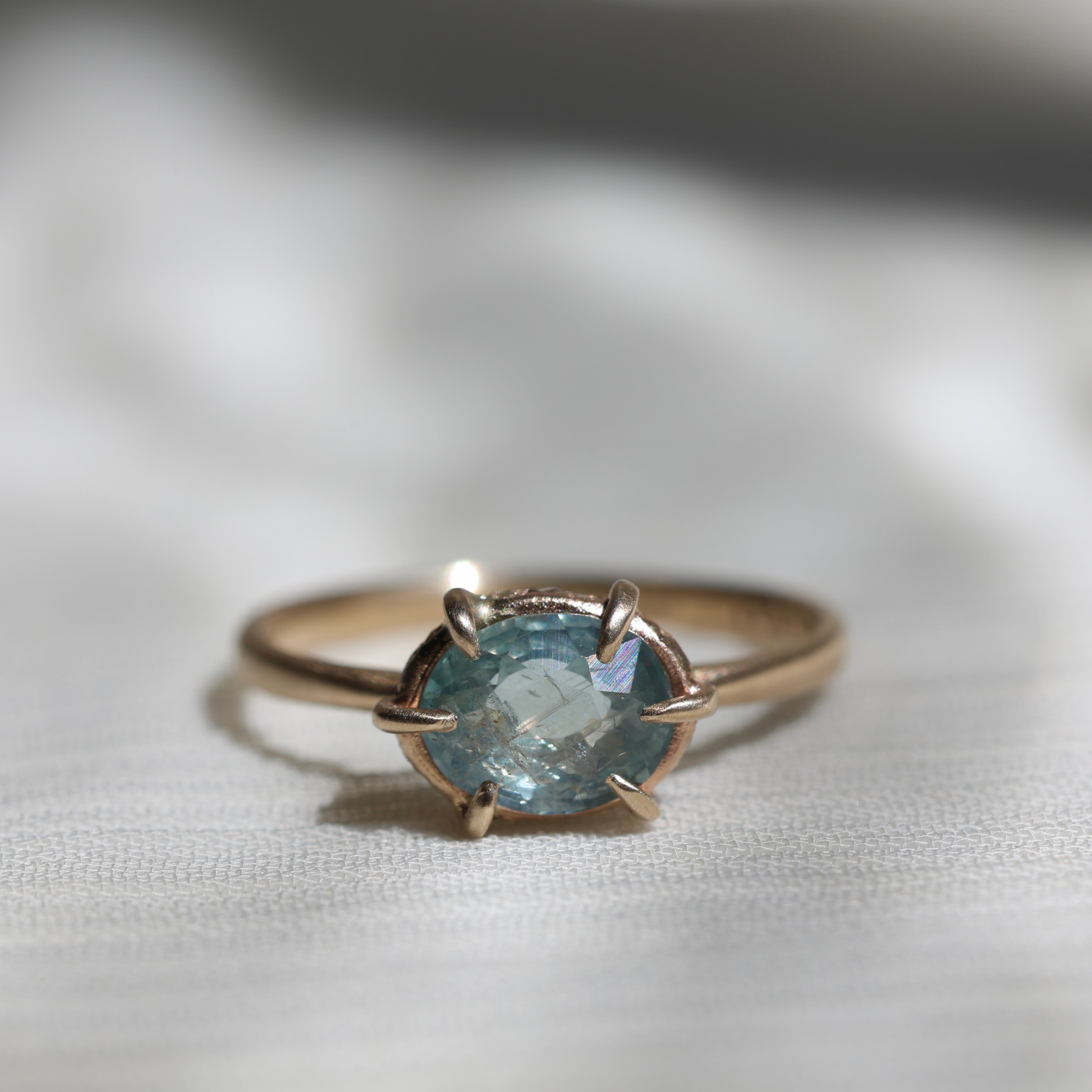 14k yellow gold ring with a light blue Montana sapphire on a soft white fabric background.
