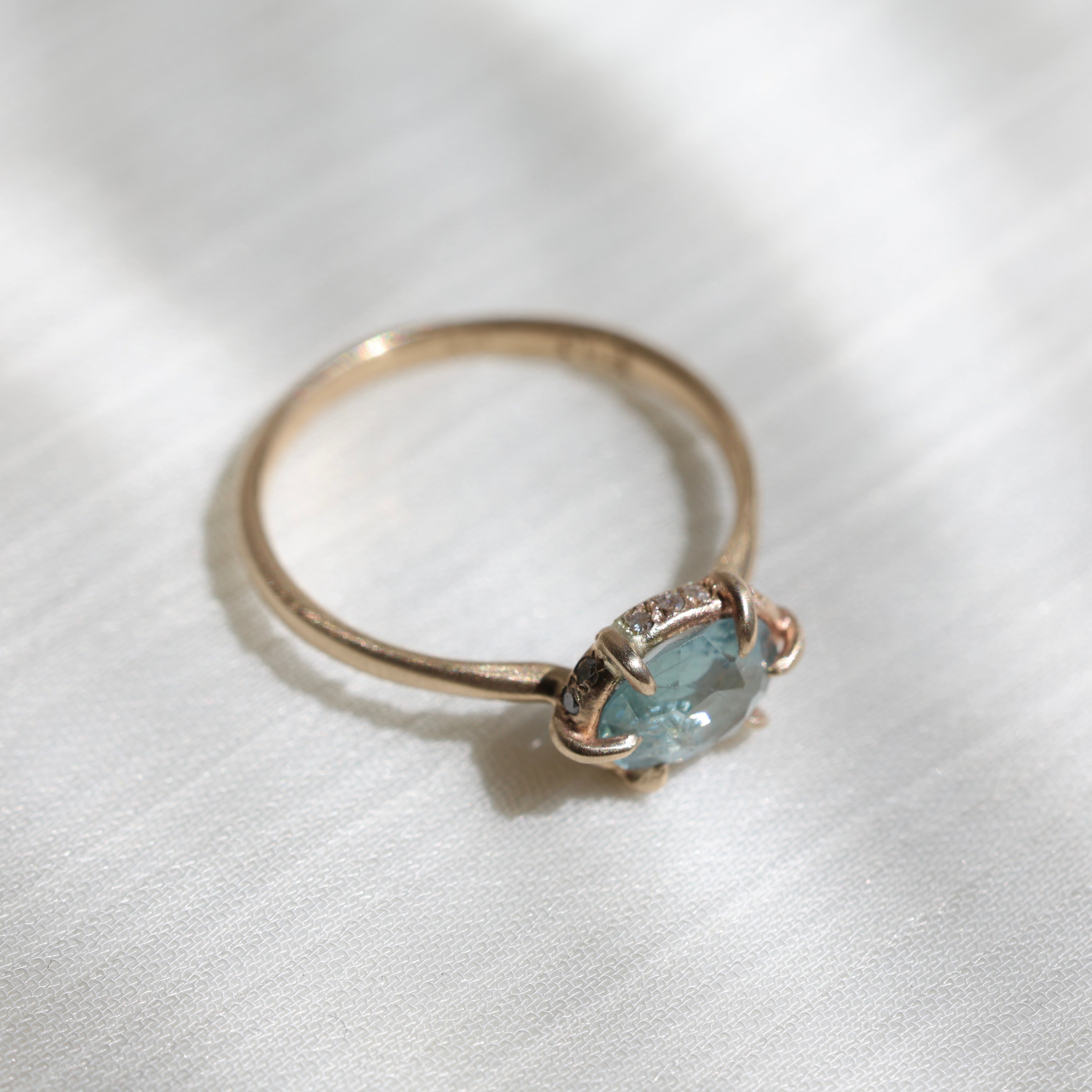 Side view of 14k yellow gold ring with a light blue Montana sapphire and a halo of small diamonds around the setting on a soft white fabric background.