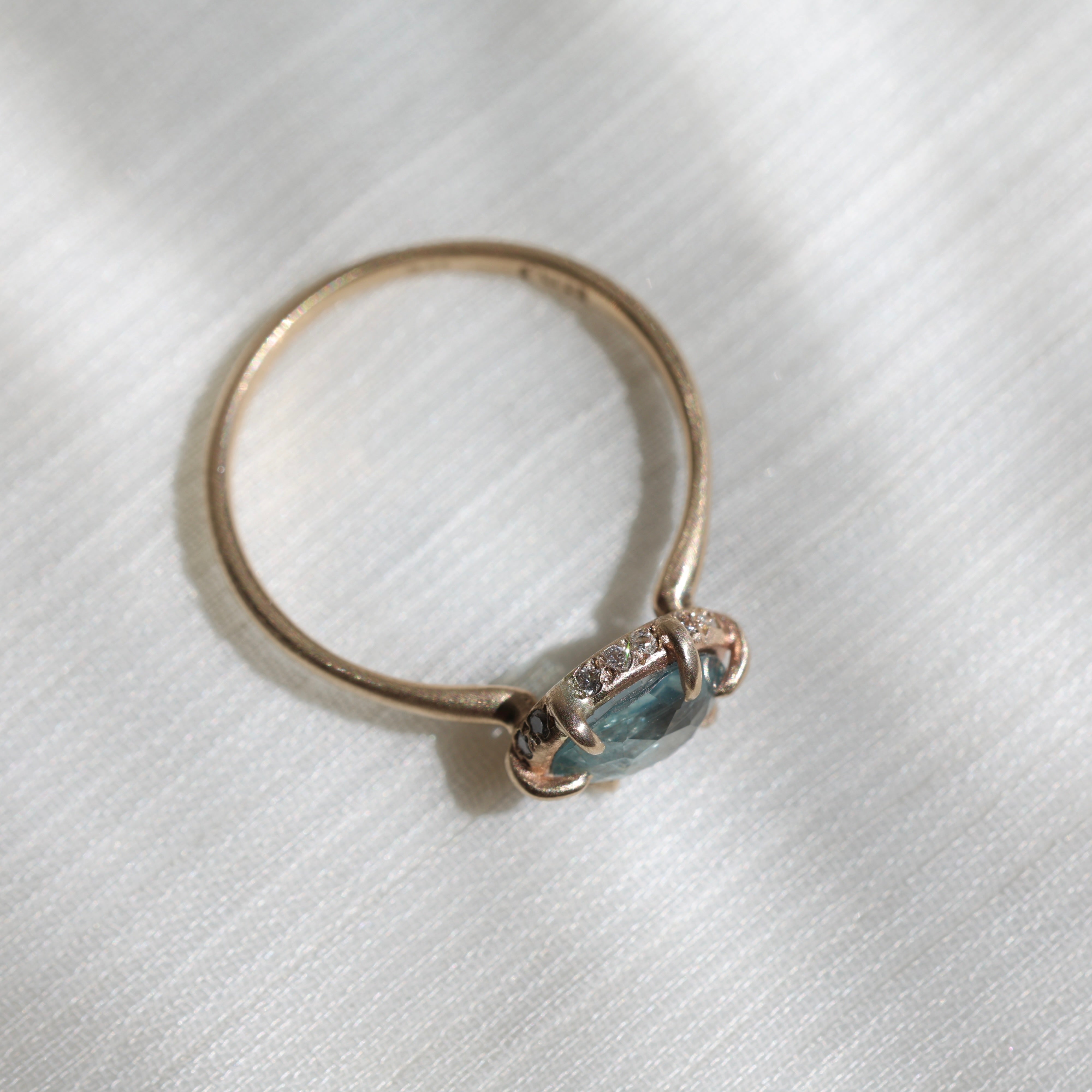 Side view of 14k yellow gold ring with a light blue Montana sapphire and a halo of small diamonds around the setting on a soft white fabric background.