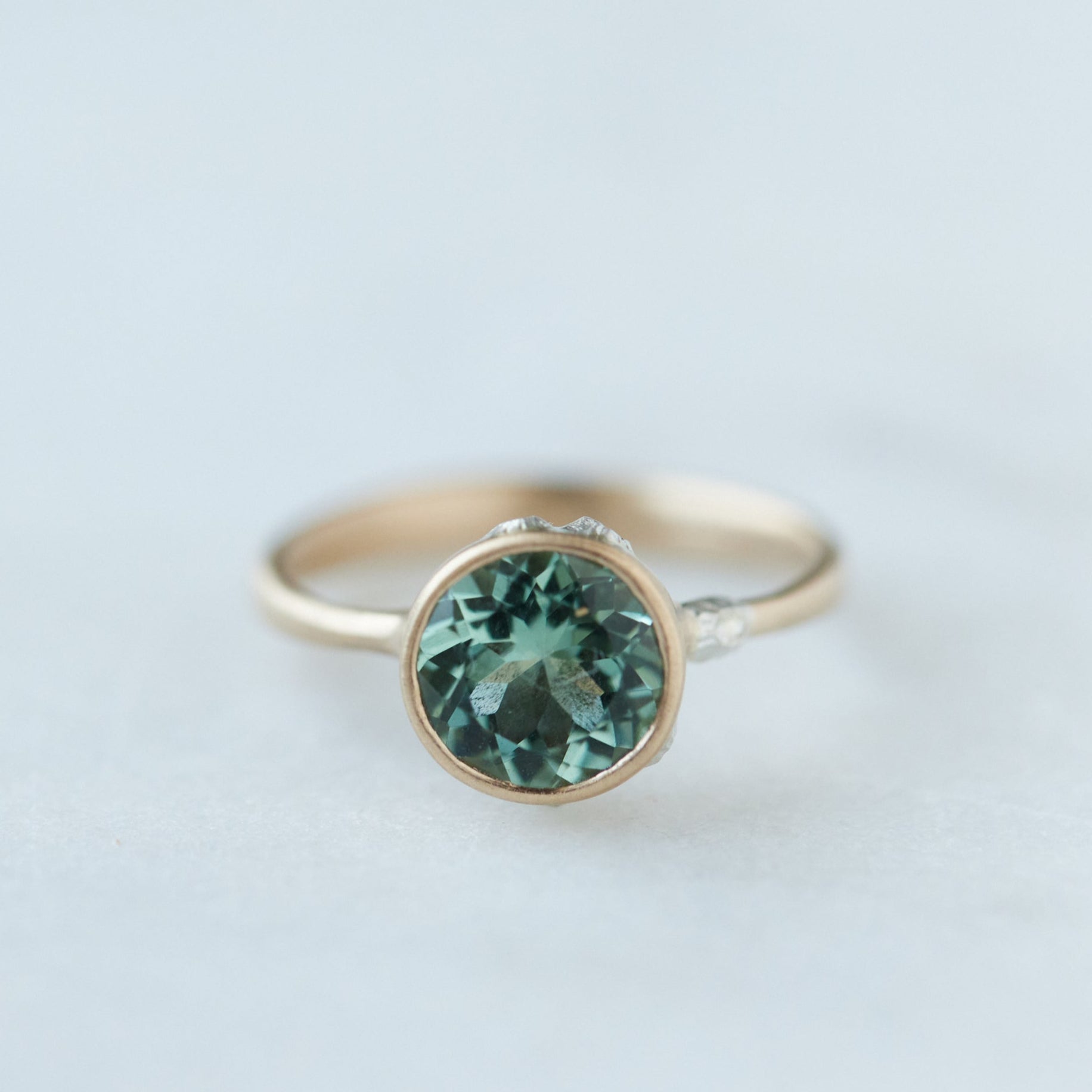 A round, icy mint tourmaline is set in a gold bezel ring with silver barnacles. Front view.