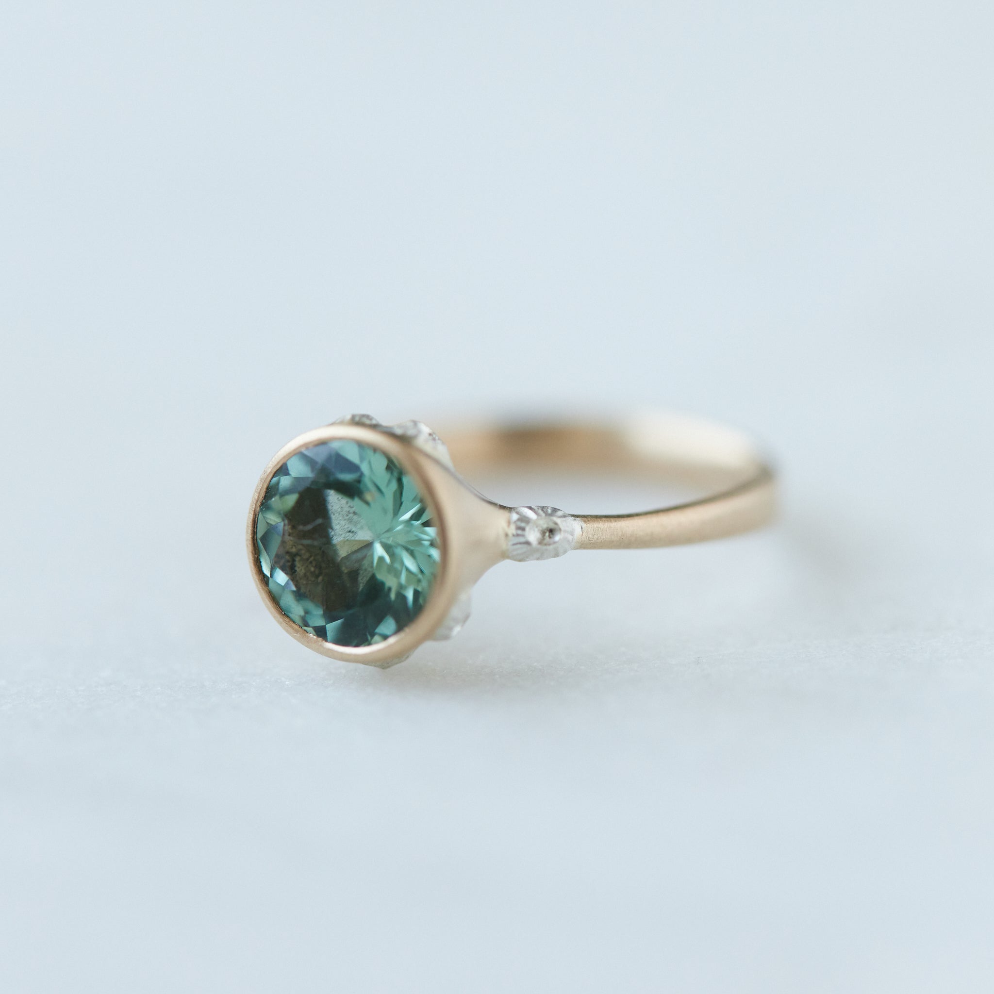 A round, icy mint tourmaline is set in a gold bezel ring with silver barnacles. Side view.