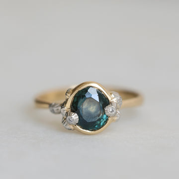 Gold ring with a teal gemstone and diamond barnacle accents on a light gray background