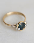 Gold ring with a teal gemstone and diamond barnacle accents on a light gray background