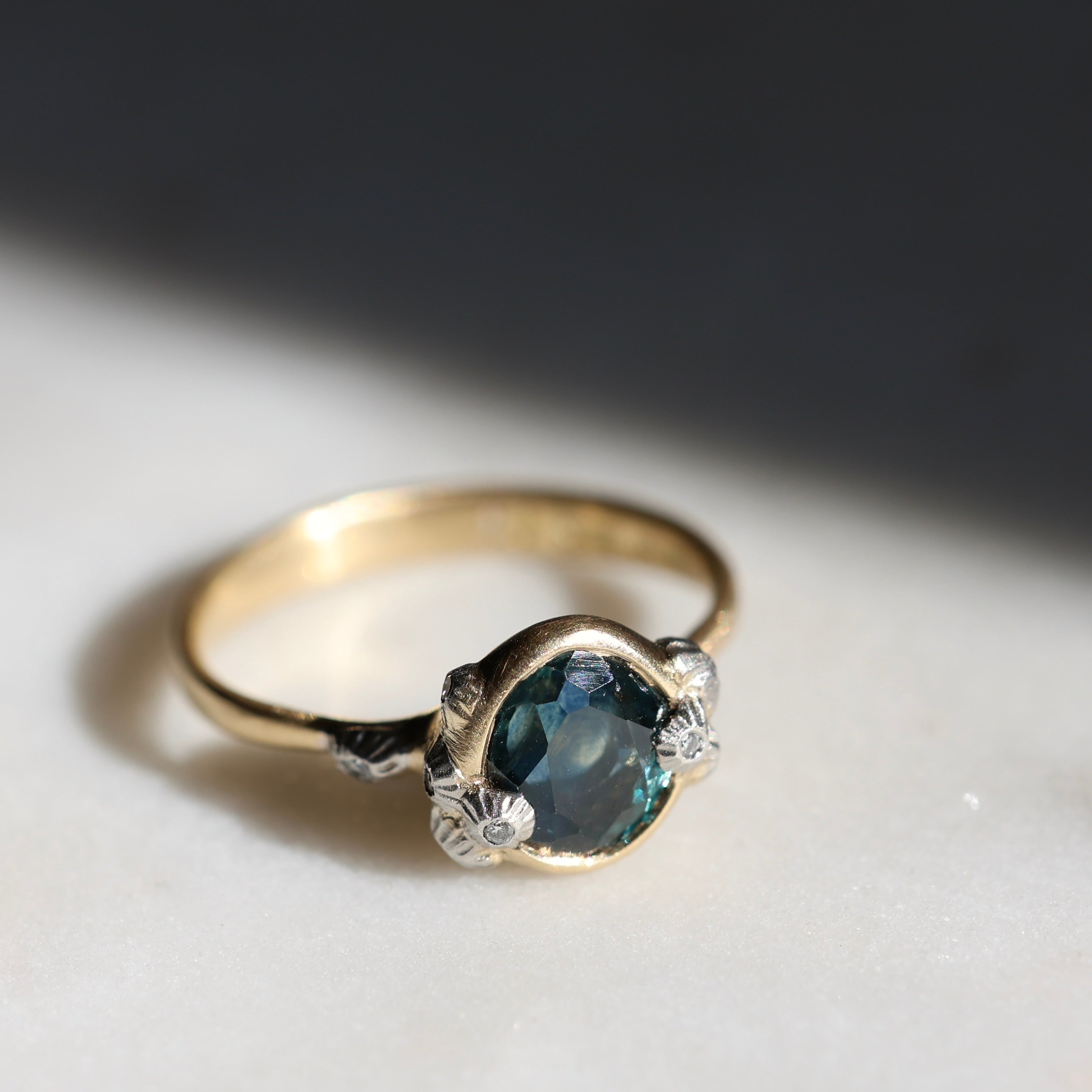 Gold ring with a teal gemstone and diamond barnacle accents on a light gray background