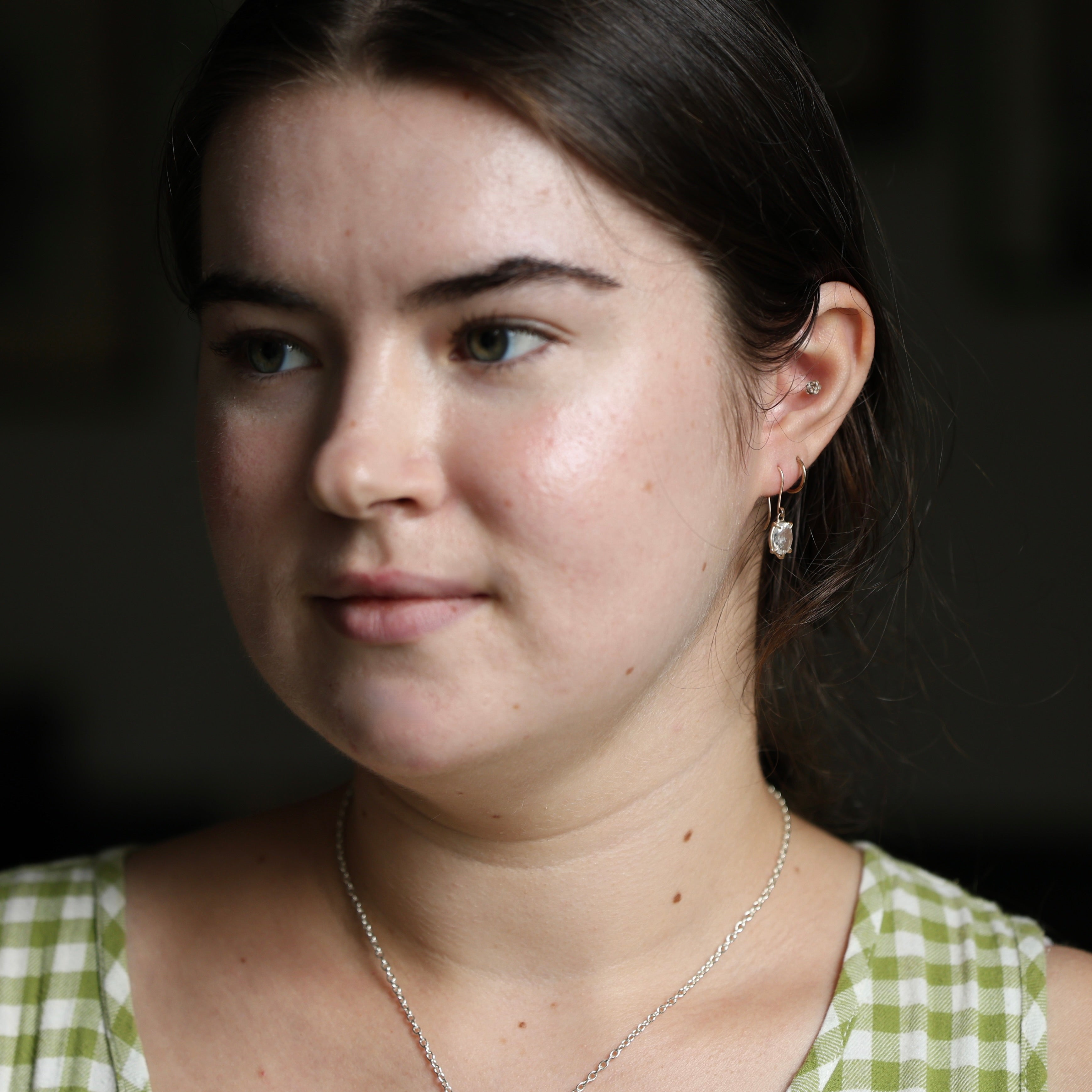 Woman wearing a green checkered dress and little white gemstone earrings with a blurred dark background