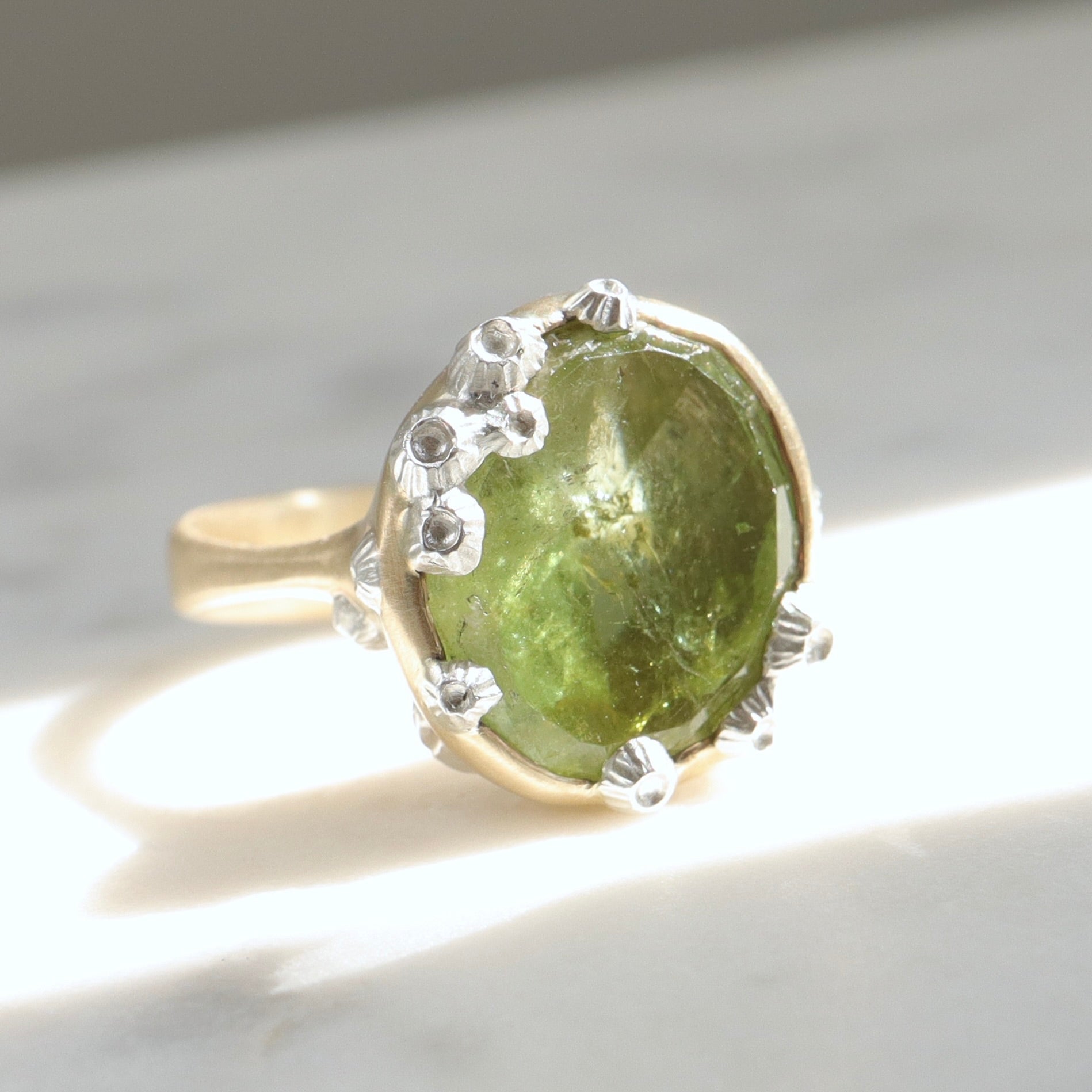 a lime green, rough-hewn tourmaline gemstone is set in a light yellow gold. Lots of little silver barnacles wrap around the setting and up over the stone. Photographed in sunlight.