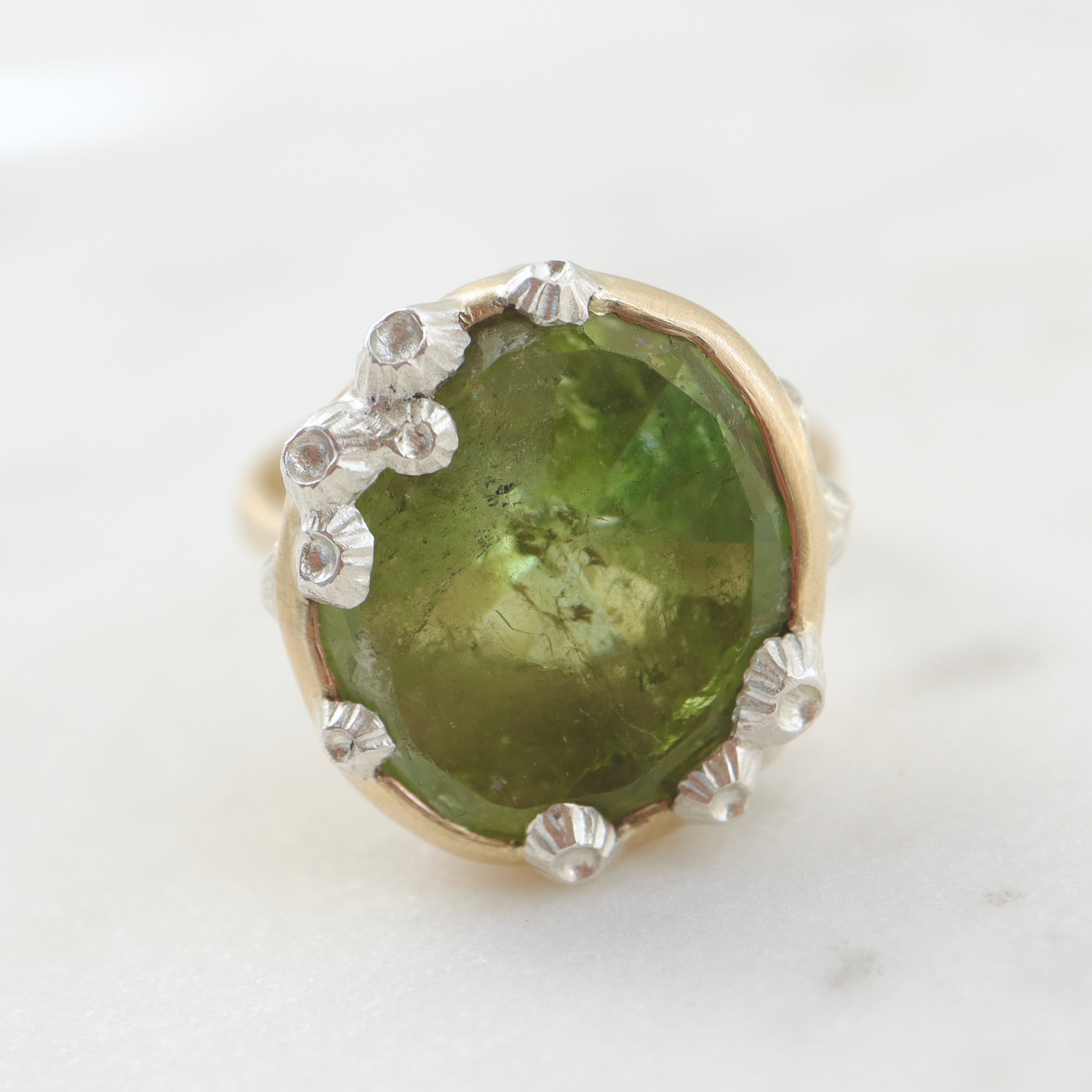 a lime green, rough-hewn tourmaline gemstone is set in a light yellow gold. Lots of little silver barnacles wrap around the setting and up over the stone. Front view.