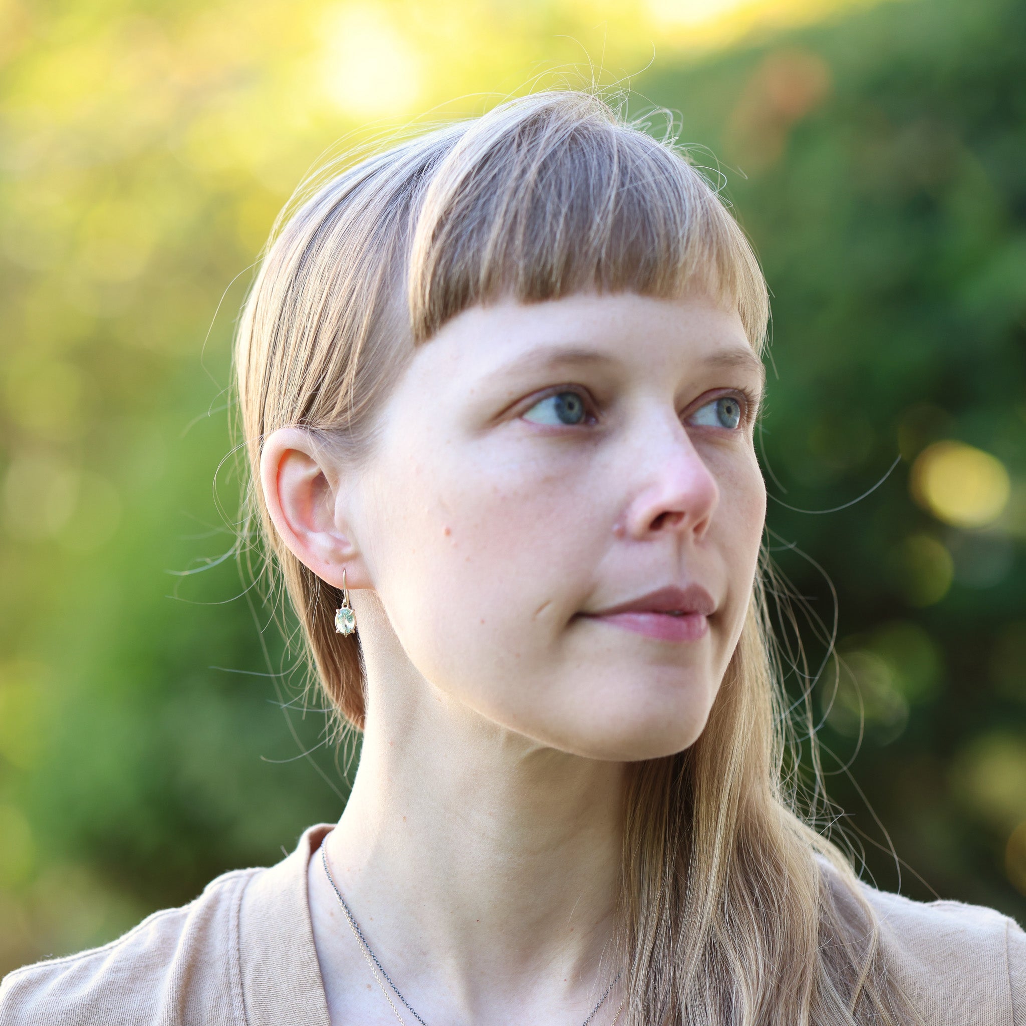 Woman with bangs wearing pale green earrings, looking to the side with a blurred green background