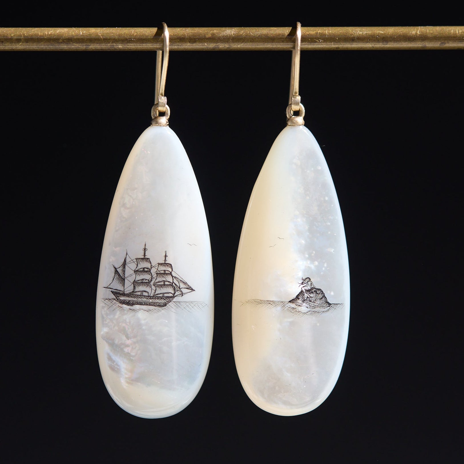 Teardrop-shaped mismatched earrings, a ship design on one and a mermaid on the other. on a black background