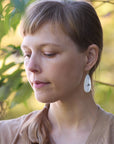 Woman with bangs wearing earrings, standing outdoors with greenery in the background