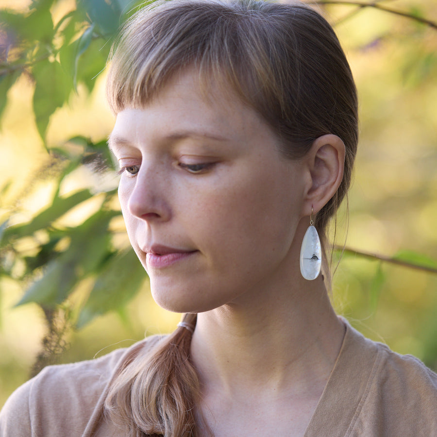 Woman with bangs wearing earrings, standing outdoors with greenery in the background