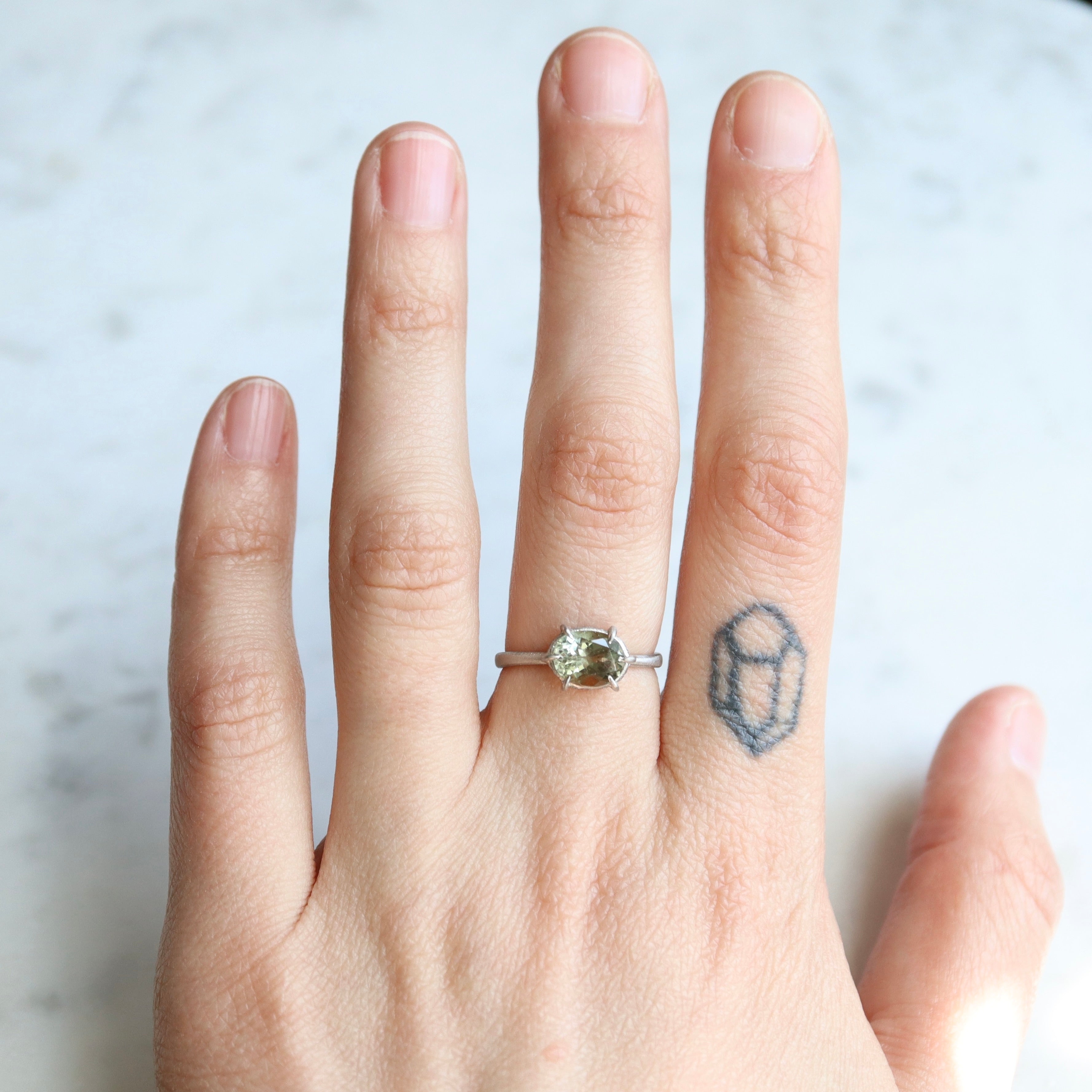 grey oval tourmaline stone set in a platinum ring with gold prongs and diamonds set around the seat. on person.