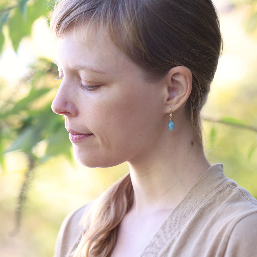 Woman with blonde hair wearing a beige top and aqua blue and gold earrings against a blurred natural background