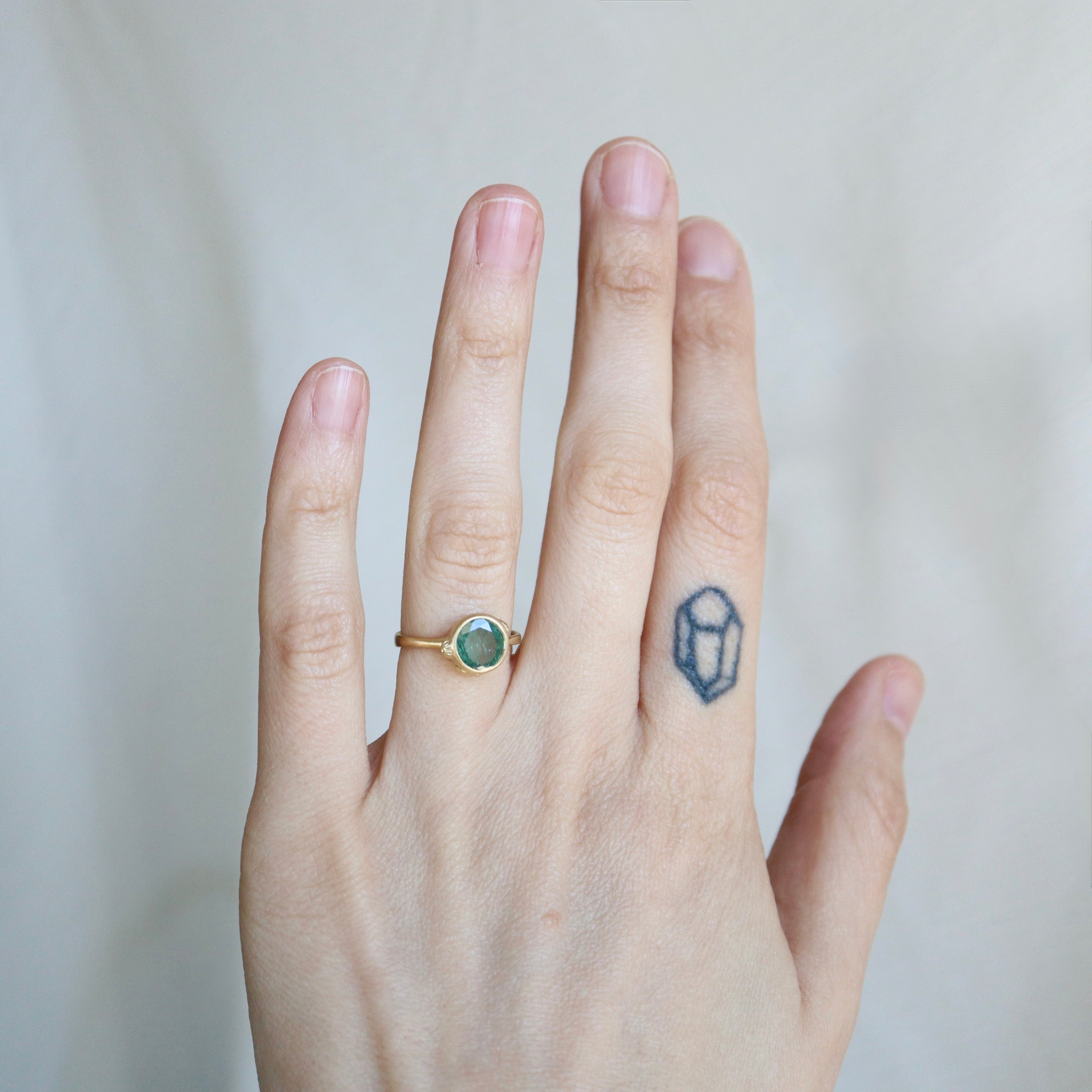 Hand wearing a gold ring with a green gemstone on a plain background