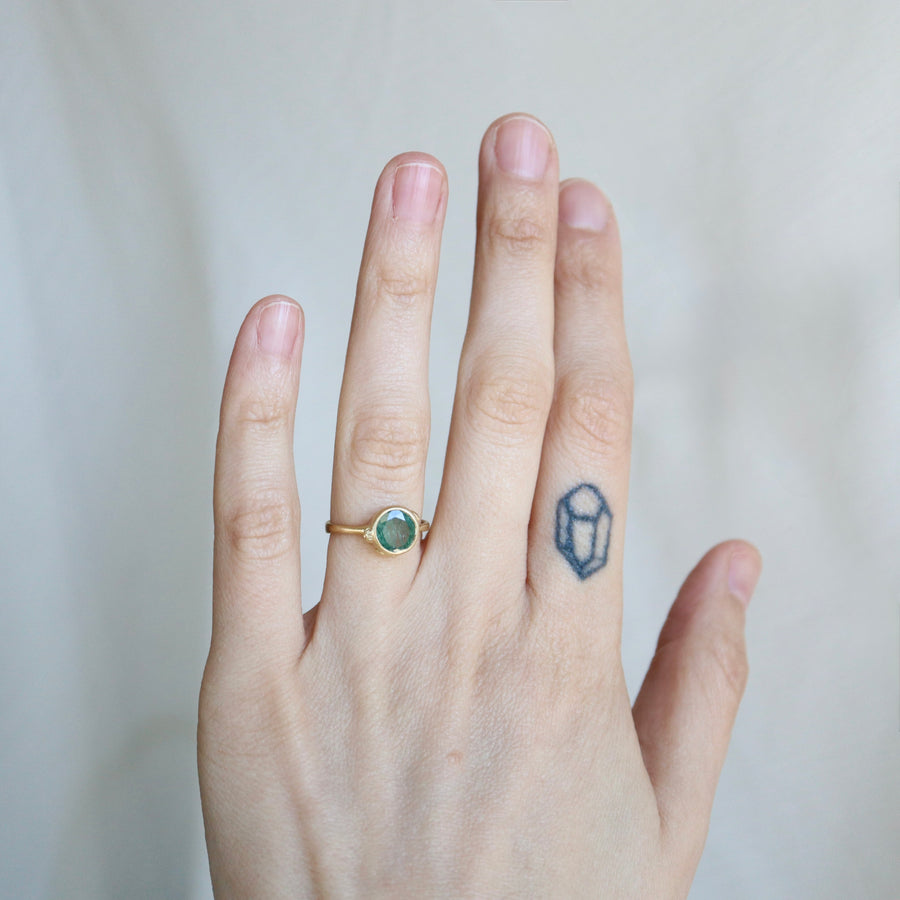 Hand wearing a gold ring with a green gemstone on a plain background