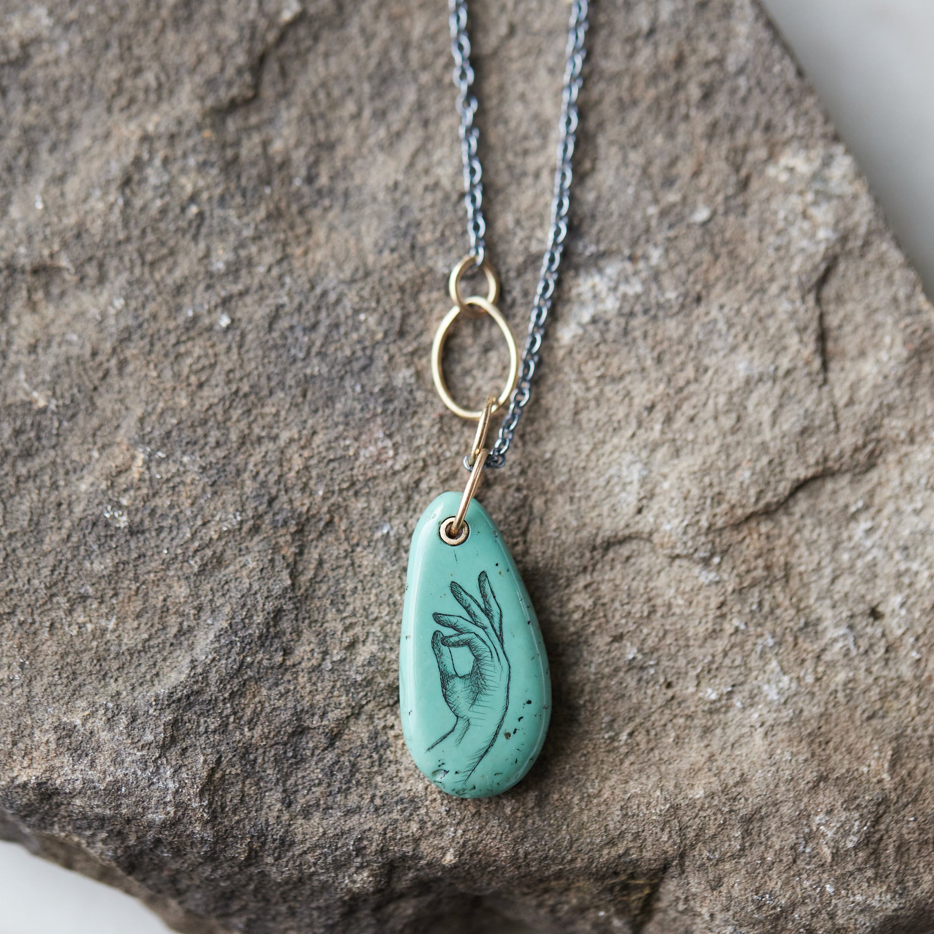 green turquoise teardrop bead with gold link and black chain. a scrimshaw hand is inked to the surface.