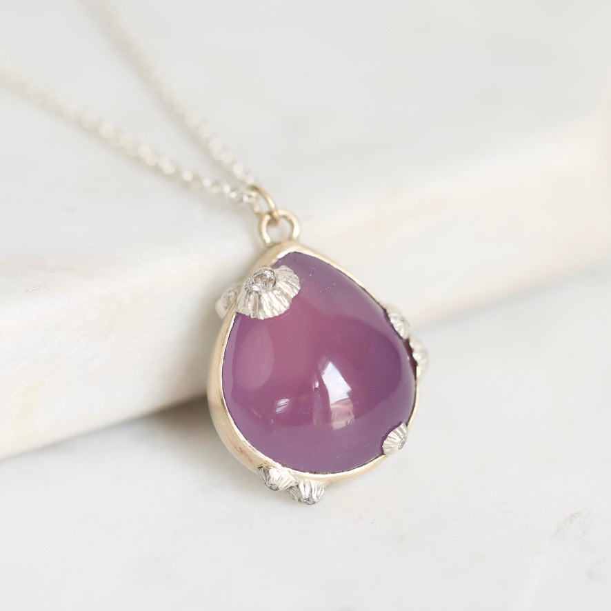 A teardrop-shaped purple gemstone pendant in a silver bezel setting with small shell-like silver accents, hanging from a silver chain on a light marble surface.