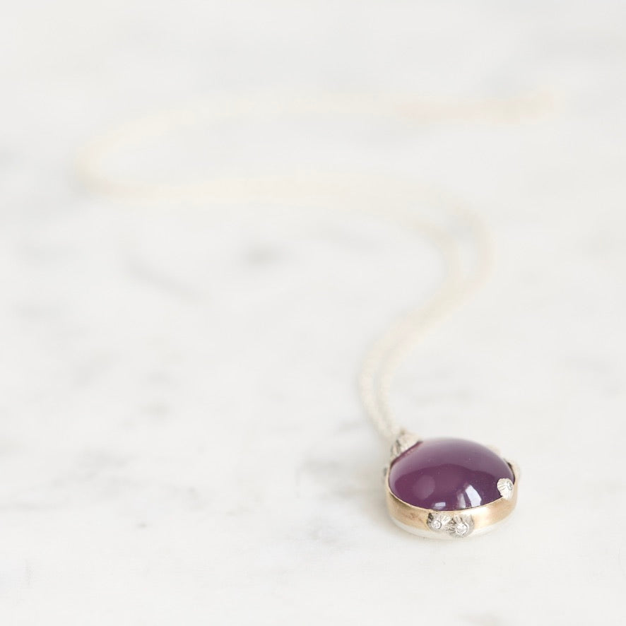 A close-up shot of a pendant necklace featuring a smooth, oval purple gemstone in a gold bezel setting with small clear accent stones, resting on a light marble surface.
