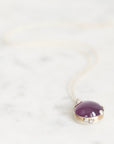 A close-up shot of a pendant necklace featuring a smooth, oval purple gemstone in a gold bezel setting with small clear accent stones, resting on a light marble surface.