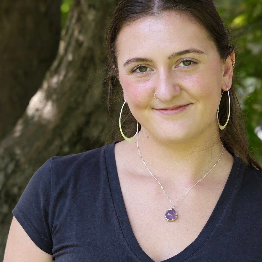 A woman with dark hair wearing a silver necklace with a purple heart pendant and large gold hoop earrings, posing outdoors in front of a tree.