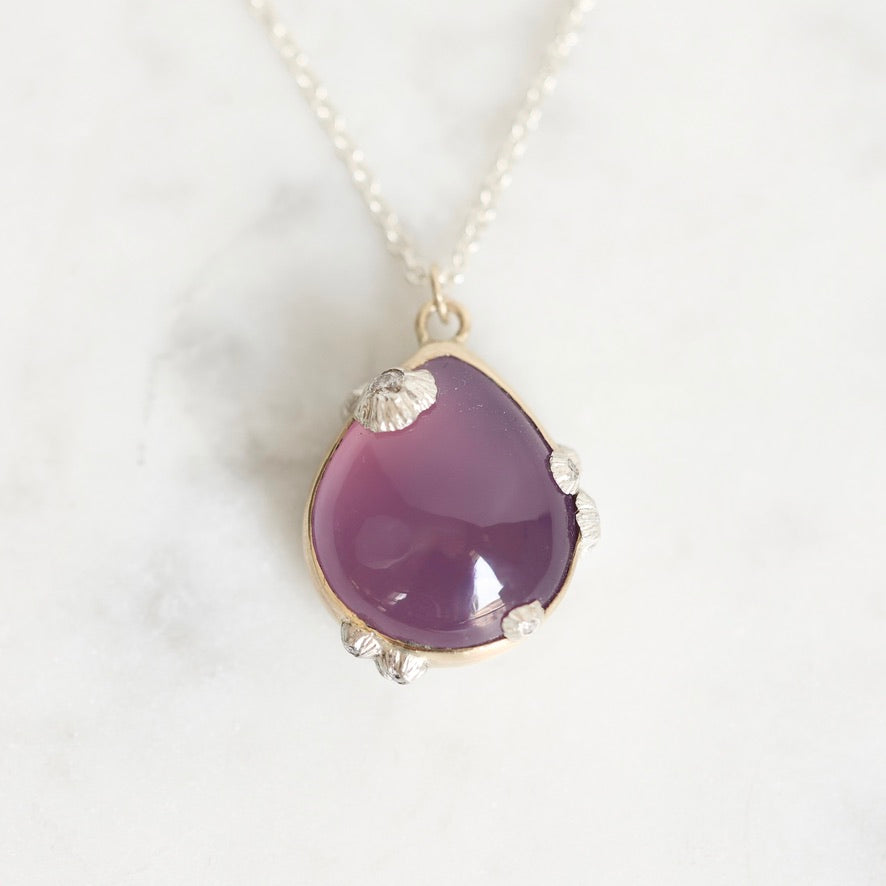 A teardrop-shaped purple gemstone pendant in a gold and silver mixed metal setting, hanging from a delicate silver chain against a light marble background.