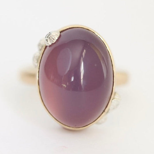 An oval cabochon gemstone ring in a gold setting with small clear accent stones on the band, shown on a plain white background.