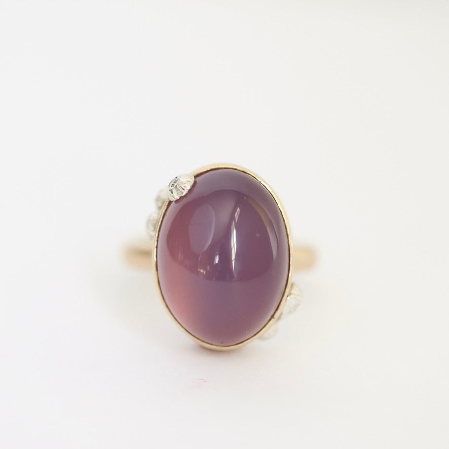 An oval cabochon gemstone ring in a gold setting with small clear accent stones on the band, shown on a plain white background.
