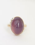 An oval cabochon gemstone ring in a gold setting with small clear accent stones on the band, shown on a plain white background.