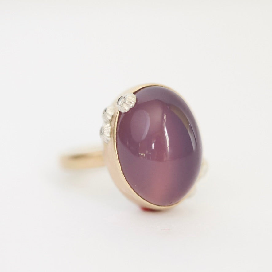 A gold ring featuring a large oval cabochon gemstone in a dusty purple shade, accented with small clear stones on the band, set against a plain white background.