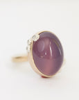 A gold ring featuring a large oval cabochon gemstone in a dusty purple shade, accented with small clear stones on the band, set against a plain white background.