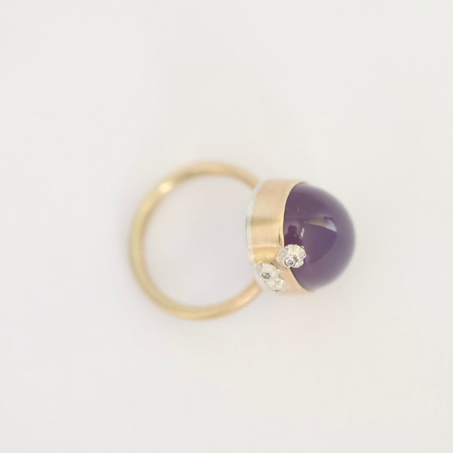 A gold ring featuring a smooth, dome-shaped dark purple amethyst stone and two small clear crystals, photographed on a clean white background.