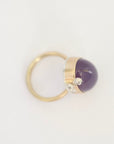 A gold ring featuring a smooth, dome-shaped dark purple amethyst stone and two small clear crystals, photographed on a clean white background.