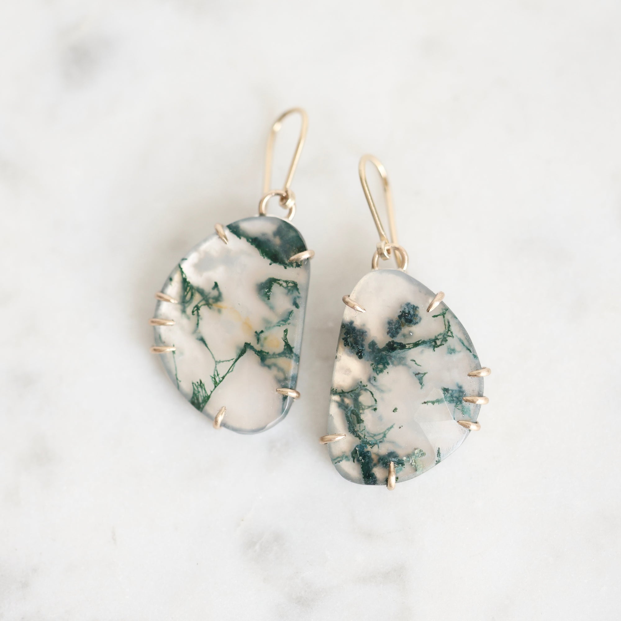 A pair of handmade teardrop-shaped dangle earrings made from white and green marbled stone, held in gold prong settings with gold fishhook ear wires, displayed on a white marble surface.