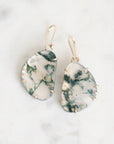 A pair of handmade teardrop-shaped dangle earrings made from white and green marbled stone, held in gold prong settings with gold fishhook ear wires, displayed on a white marble surface.