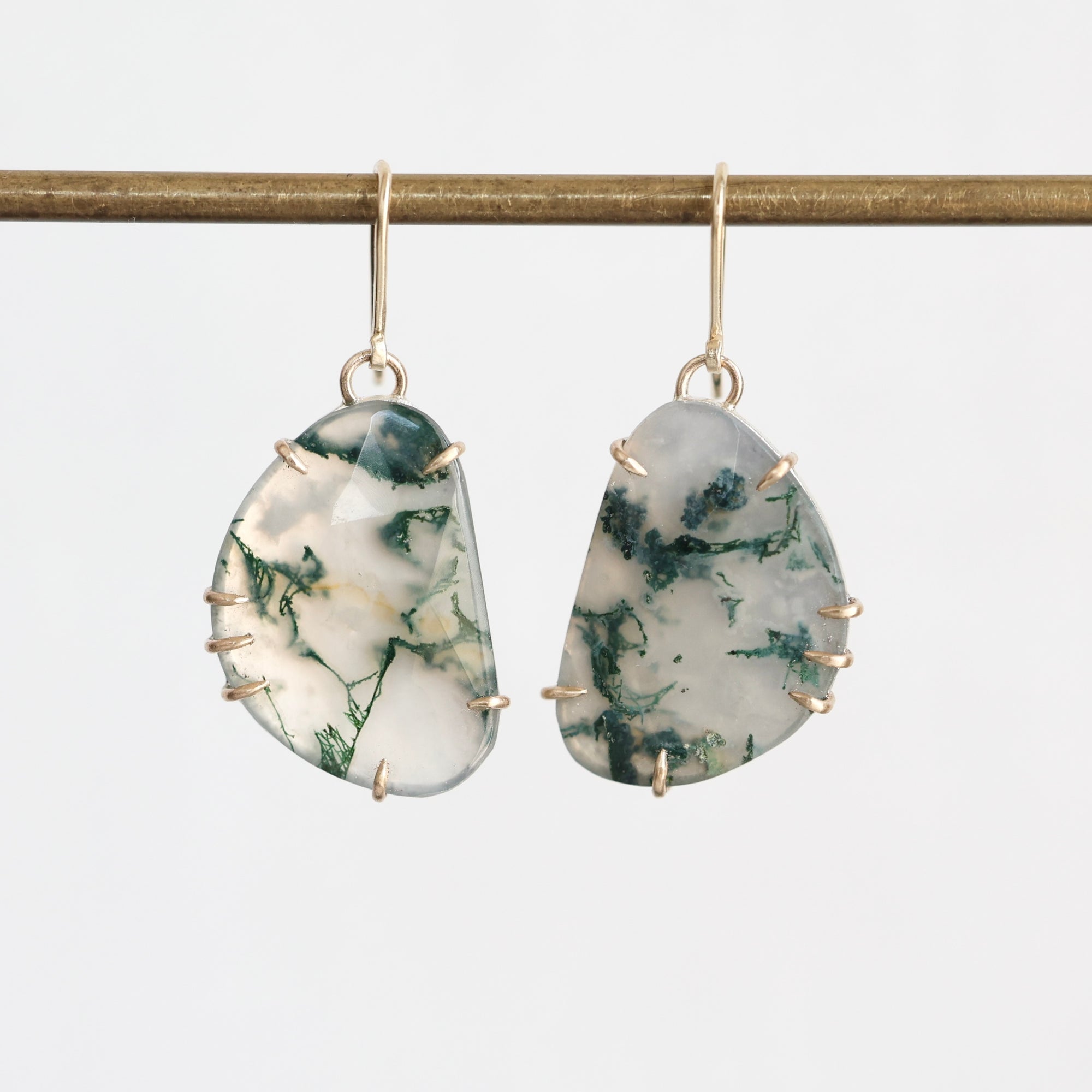 A pair of dangle earrings with irregular organic-shaped white and green marbled stone pendants in gold bezel settings, hanging from gold fishhook wires against a light gray background.