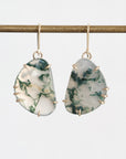 A pair of dangle earrings with irregular organic-shaped white and green marbled stone pendants in gold bezel settings, hanging from gold fishhook wires against a light gray background.