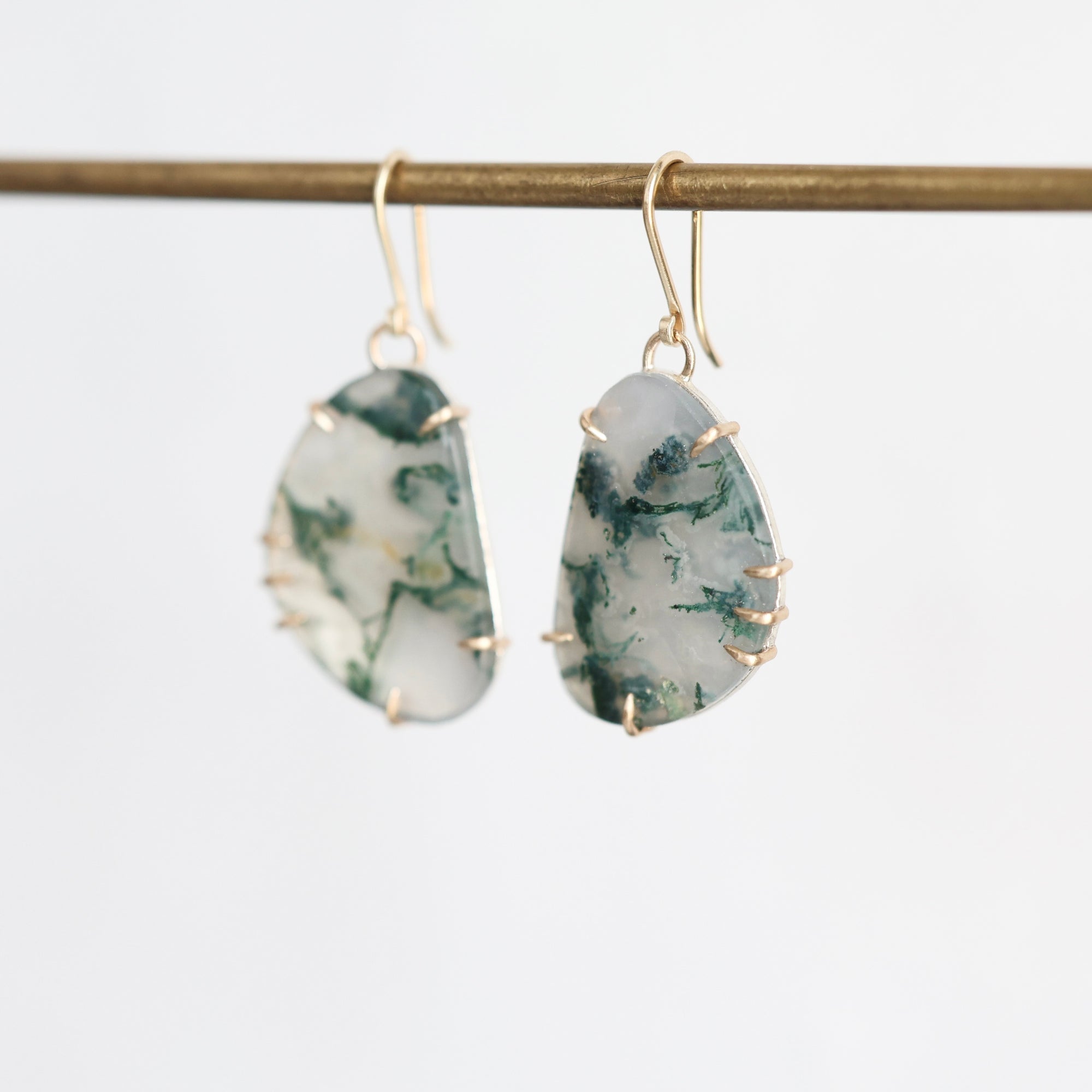 A pair of gold-tone dangle earrings with irregular, organic-shaped green and white marbled stones, hanging from a gold-colored horizontal bar against a clean white background.