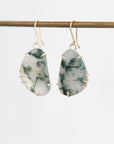 A pair of gold-tone dangle earrings with irregular, organic-shaped green and white marbled stones, hanging from a gold-colored horizontal bar against a clean white background.