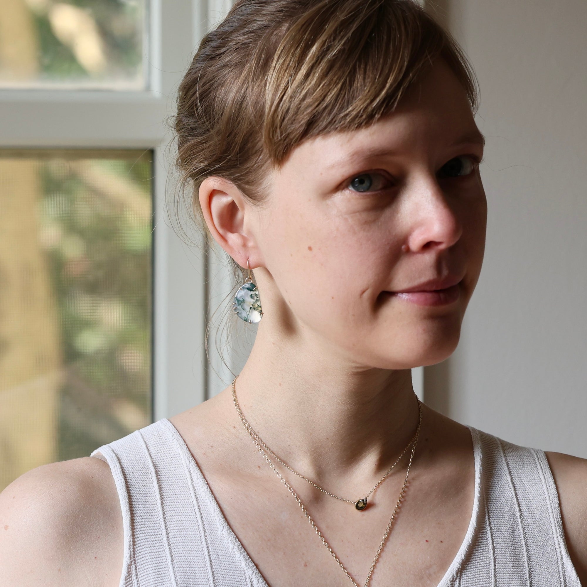 A woman with light brown hair pulled back, wearing a white ribbed tank top, layered gold necklaces, and a dangling silver-toned earring with a clear, textured stone, photographed in soft natural light near a window.