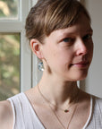 A woman with light brown hair pulled back, wearing a white ribbed tank top, layered gold necklaces, and a dangling silver-toned earring with a clear, textured stone, photographed in soft natural light near a window.