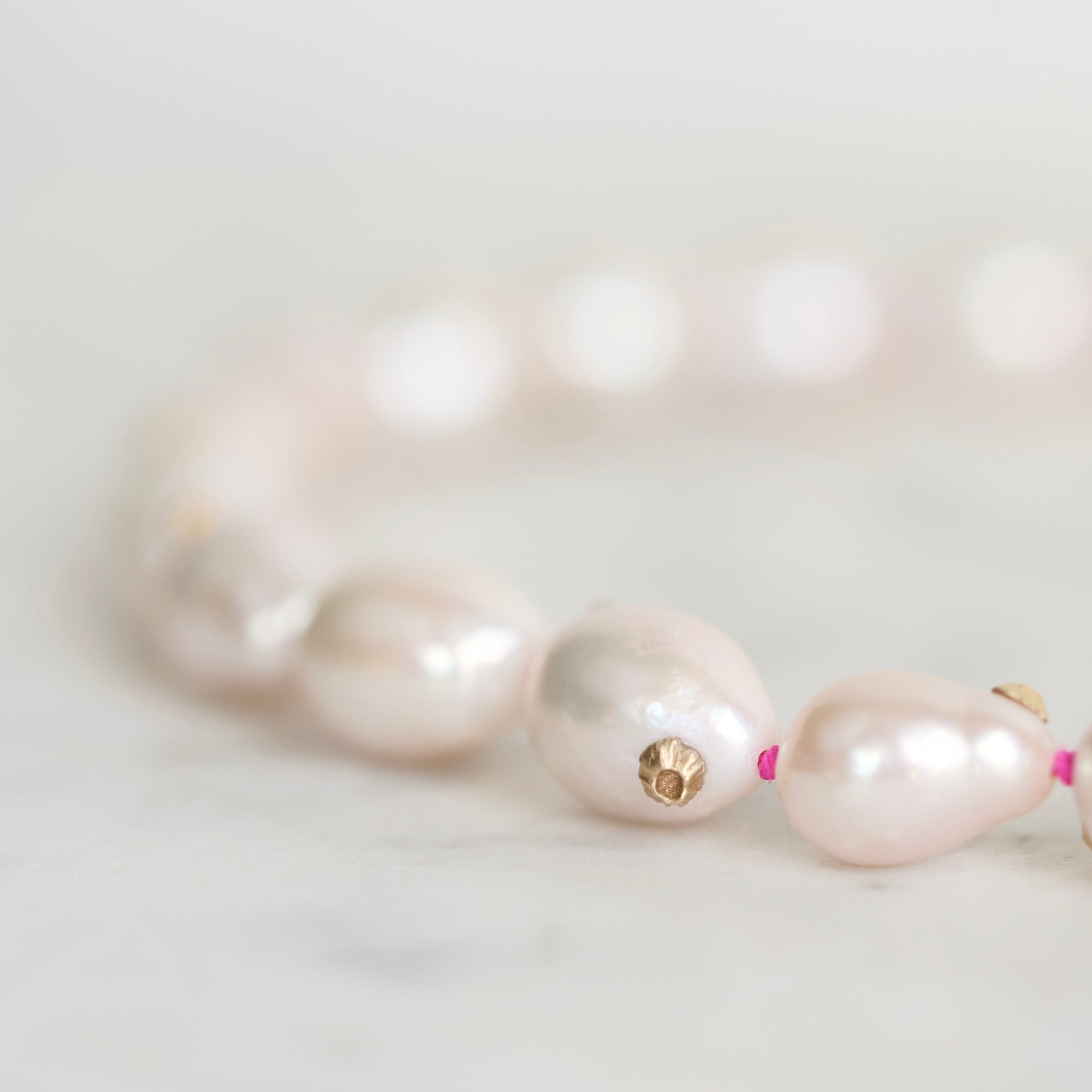A close-up of a pearl beaded bracelet on a light marble surface. The bracelet features irregular, lustrous pale pink pearls strung with small bright pink beads and a small gold flower-shaped charm in the center.