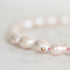 A close-up of a pearl beaded bracelet on a light marble surface. The bracelet features irregular, lustrous pale pink pearls strung with small bright pink beads and a small gold flower-shaped charm in the center.