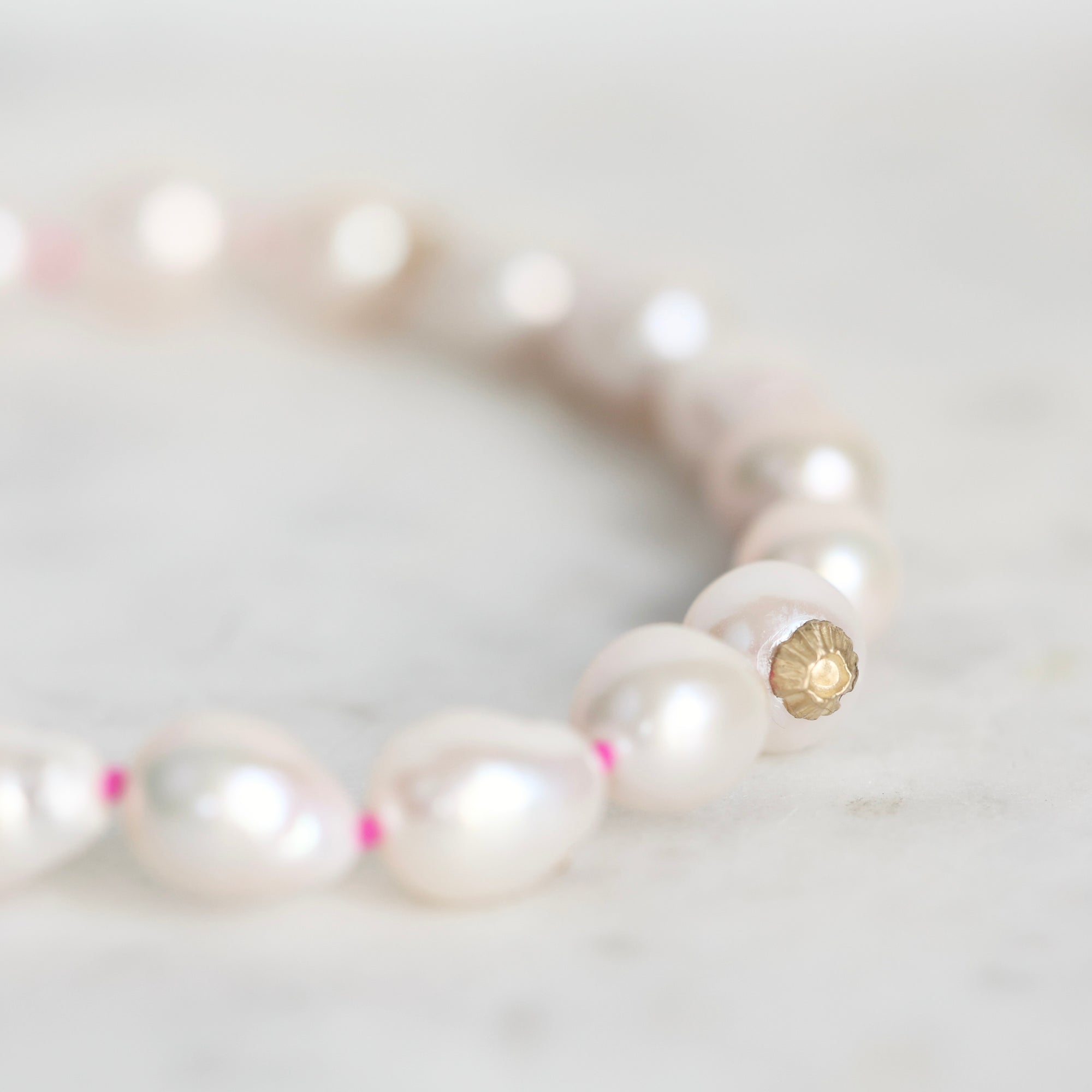 A close-up of a beaded bracelet featuring irregular white freshwater pearls separated by tiny pink spacers, with a gold-toned flower-shaped bead near the front, resting on a light marble surface.