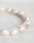 A close-up of a beaded bracelet featuring irregular white freshwater pearls separated by tiny pink spacers, with a gold-toned flower-shaped bead near the front, resting on a light marble surface.